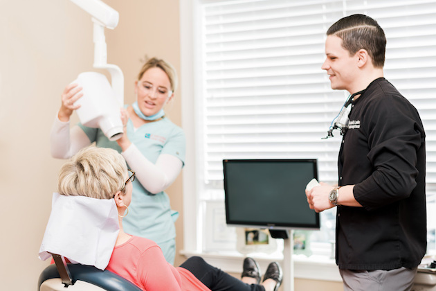 dentist smiling at righ with hygienist in background with x-ray machine and happy patient looking at them both